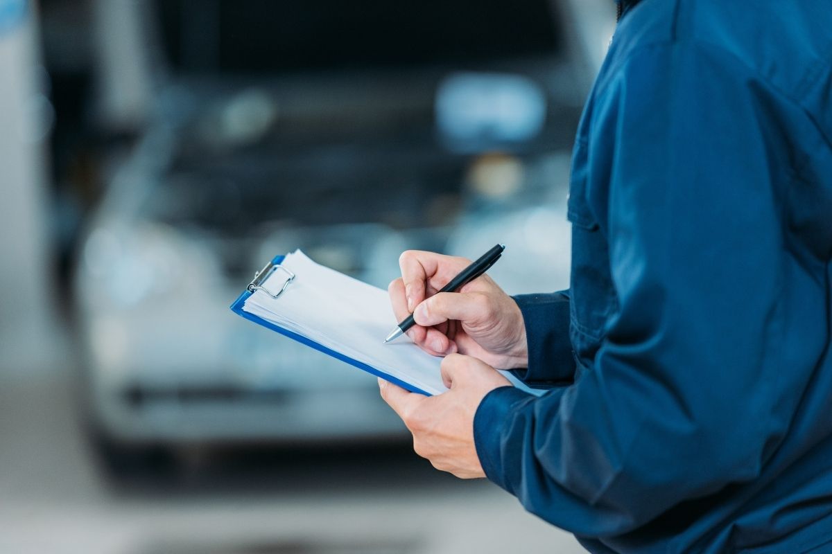 prikaz majstora koji popunjava servisnu knjižicu, u pozadini automobil bele boje sa otvorenom haubom